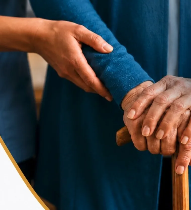A caregiver's hands resting on an elderly person's hands on a walking stick